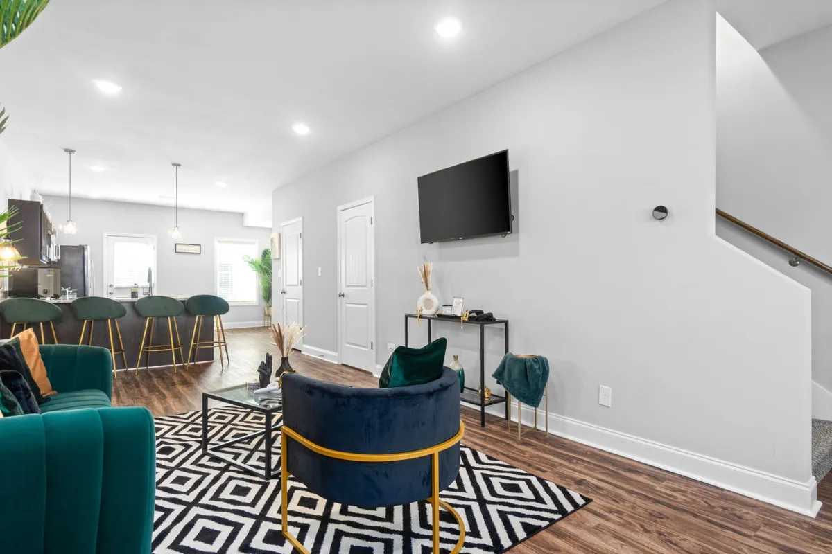 This inviting living area features a plush green velvet sofa and a stylish blue armchair, anchored by a bold black and white patterned rug. The open-concept layout seamlessly connects the cozy lounge space to a modern kitchen bar with sleek stools, while a wall-mounted TV and thoughtful decor create the perfect setting for relaxing evenings, movie nights, or gathering with friends.