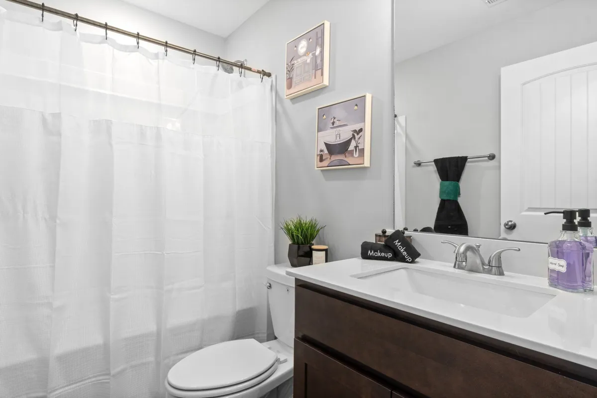 A bright, inviting bathroom with a white shower curtain, modern vanity with sink, makeup organizer, and framed artwork. The atmosphere is serene and welcoming, highlighting attention to detail and personal care.