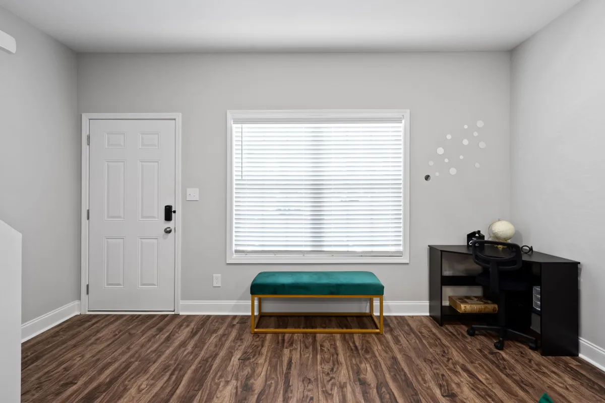 A tranquil workspace featuring a modern black desk beside a large window with blinds, a green upholstered bench, and abundant natural light. The setting is ideal for focused work, reading, or taking a quiet moment of inspiration.