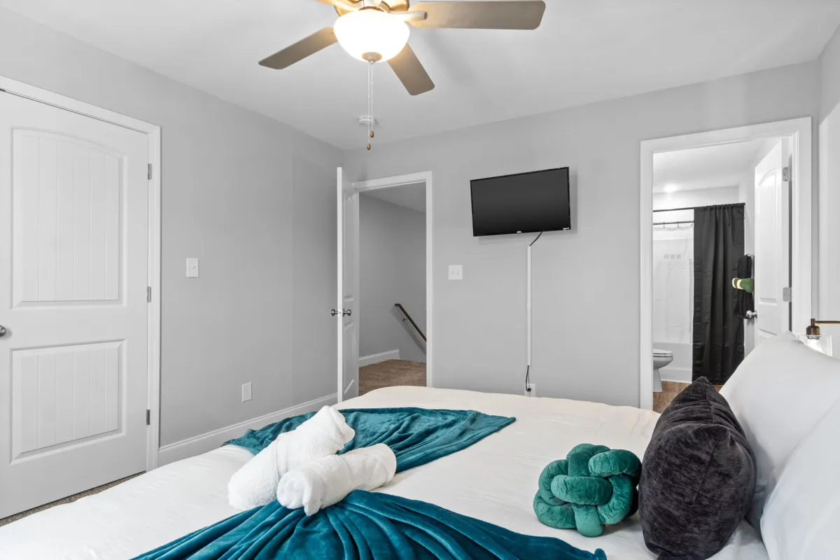 A serene bedroom with a bed dressed in a teal throw, folded towels neatly placed, and a wall-mounted TV. The tranquil mood, accentuated by calm colors and soft lighting, highlights the readiness for guests with fresh linens and towels.